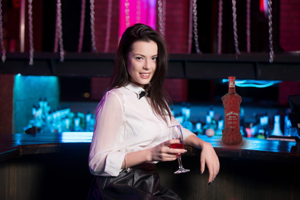 Attractive smiling young brunette woman wearing white shirt and black bow tie sitting at bar counter, holding glass of pink cocktail